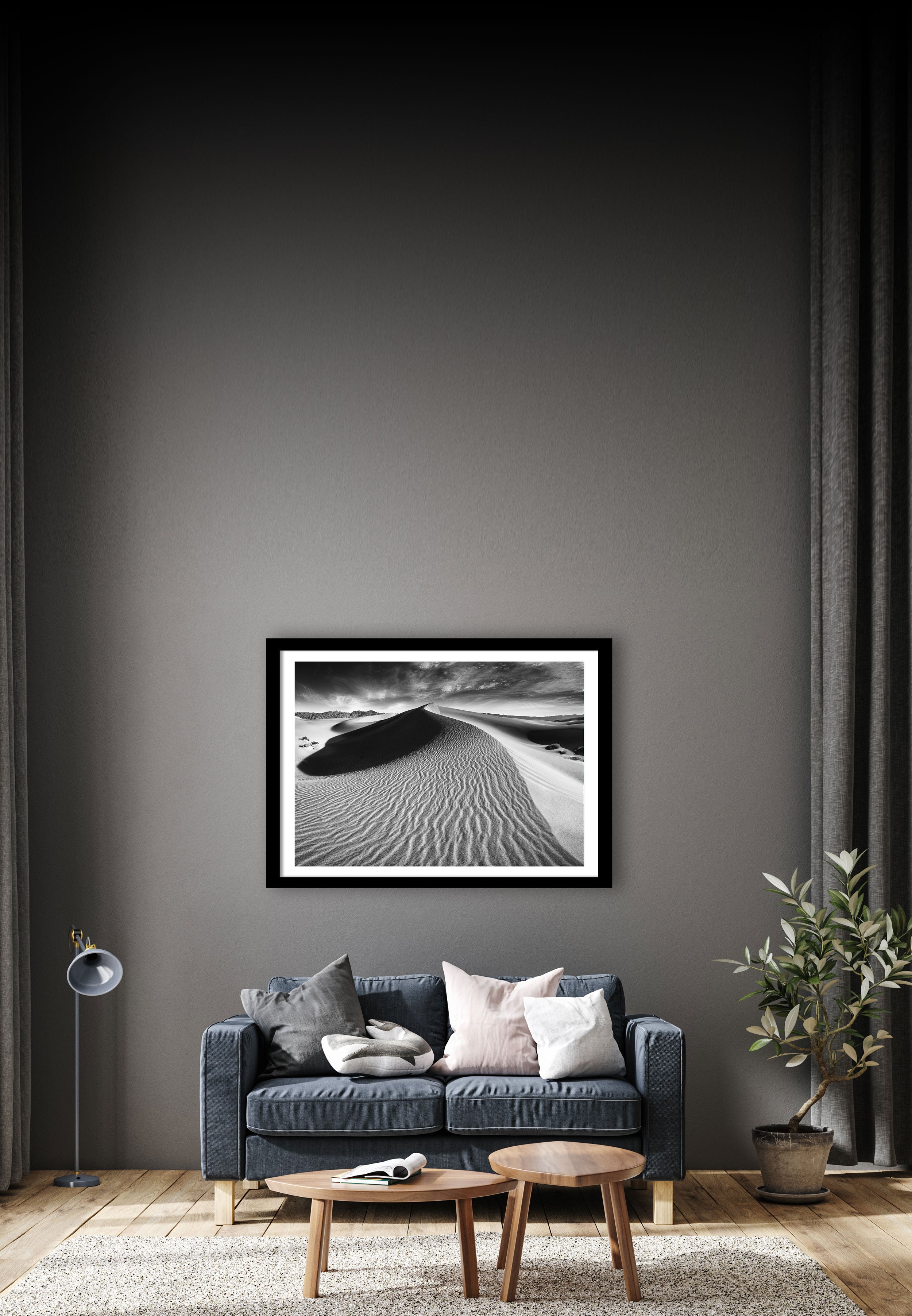 A minimalist living room features a dark gray accent wall with a framed black and white photograph of a desert dune. A dark gray sofa with throw pillows sits below, accompanied by a wooden coffee table, a potted plant, a floor lamp, and a beige rug.